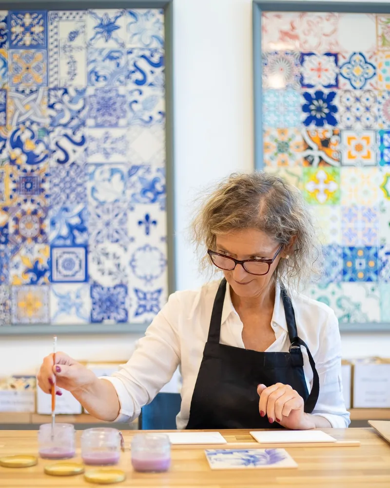 Persona con delantal pintando un azulejo con pincel fino en un taller, con paneles de azulejos de motivos florales al fondo
