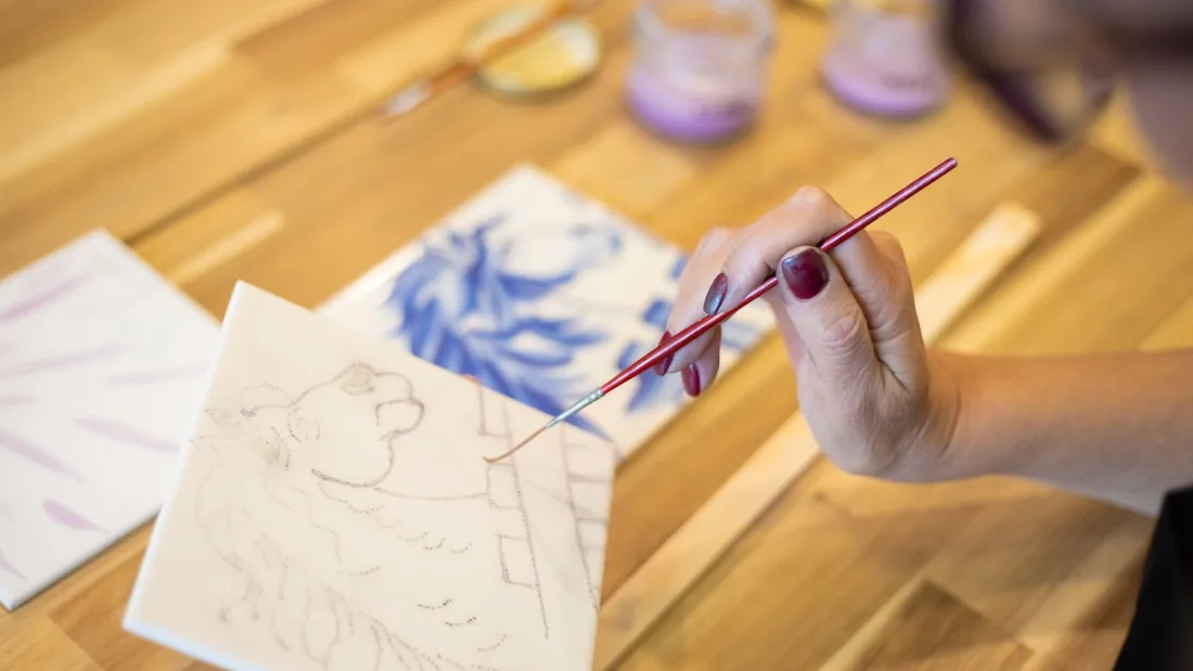 Mano sosteniendo un pincel fino sobre un azulejo blanco con el dibujo trazado a lápiz, junto a otra pieza pintada en azul